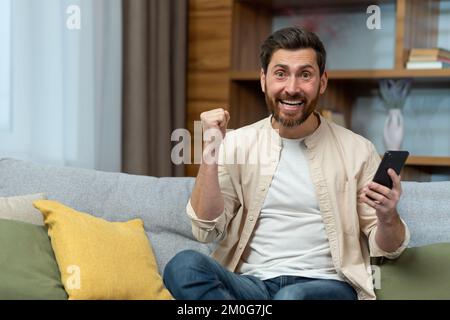 Portrait of man at home happy and celebrating online casino victory, rejoices satisfied with achievement and looks at camera holding smartphone and hand raised in triumph gesture. Stock Photo