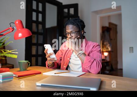 Worried young African man sitting at desk with laptop checking phone, receiving bad news via sms Stock Photo