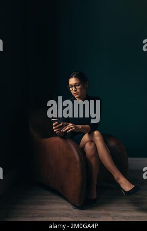 Sophisticated woman using a mobile phone while sitting on an armchair in her office. Modern woman reading a text message on her smartphone. Stock Photo