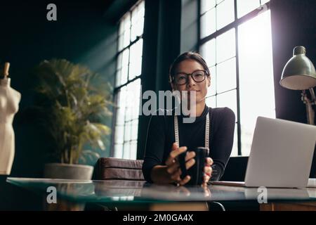 Asian woman holding cup of coffee and looking at camera. Female designer sitting relaxed at her desk with coffee. Stock Photo