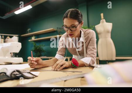 Young asian woman working in her workshop and making a design draft on paper. Female fashion designer designing new dress pattern on paper. Stock Photo