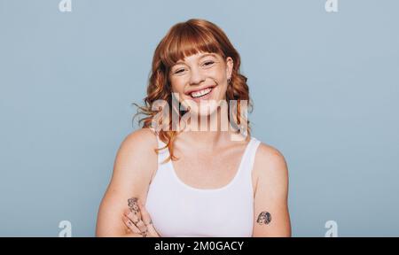 Happy red haired woman smiling at the camera in a studio. Portrait of a young caucasian woman standing in a white tank top. Stock Photo