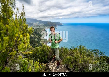 Hiker standing on a rock, view of steep wooded coast and sea, in the background village Porto da Cruz and eagle rock Penha de Aguia, coastal landscape Stock Photo