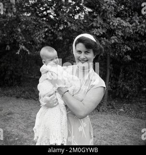 1950s, historical, christening, standing outside for a photo, a happy mother holding her just christened child which is wrapped in a crochet patterned baby shawl, with a sweet smile on its little face, England, UK. Stock Photo