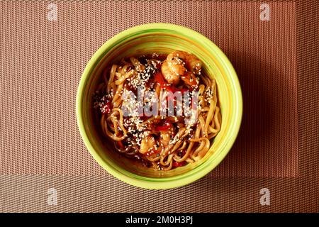 Udon stir fry noodles with vegetables, shrimps and sesame seeds in green plate bowl on noisy brown table mat background. Top view, close-up Stock Photo