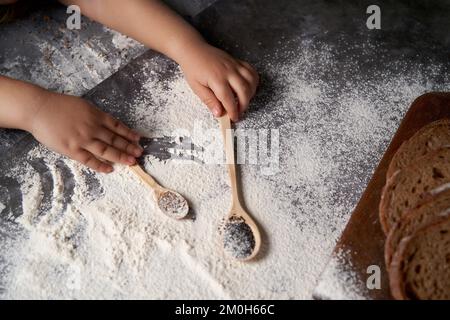 The hands of a kid who helps to make pastries . The child holds wooden spoons with seeds, flour is scattered on the table. High angle. Stock Photo