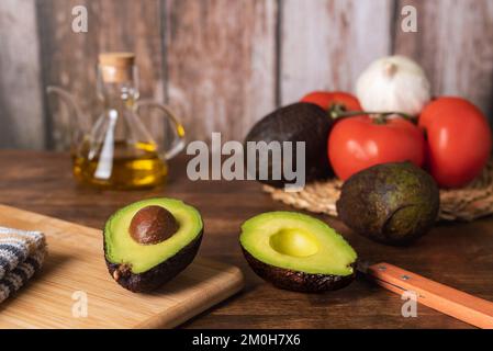 Avocado cut in two, with a rustic background with tomatoes, garlic, more avocados and oil. Stock Photo