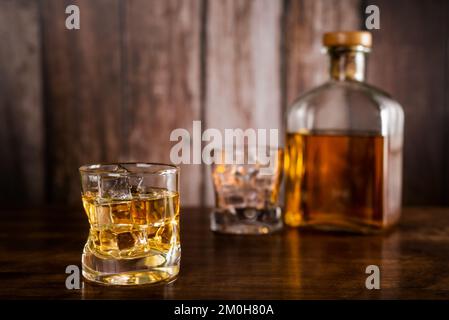 A glass of whiskey with ice cubes, with a bottle of whiskey and an empty glass in the background out of focus. Stock Photo
