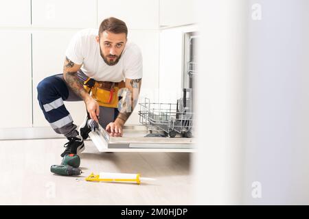 Young modern serviceman in worker suit during the repairing of the dishwasher on the domestic kitchen Stock Photo