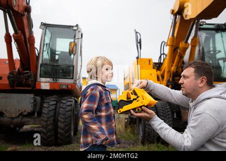Dad and son, against background of construction equipment - excavators, are enthusiastically examining a toy bulldozer, which a man holds in his hands Stock Photo