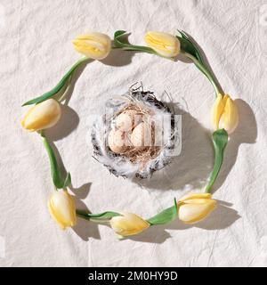 Easter background with yellow tulips and beige eggs in nest on off white textile. Top view, flat lay, simple, minimal arrangement Stock Photo