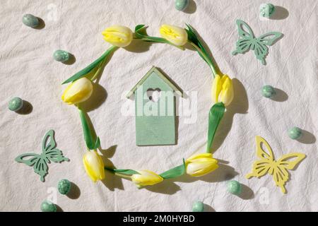Easter background with yellow tulips around decorative bird house. Textile background, flat lay with mint green quail eggs and yellow wood butterflies Stock Photo