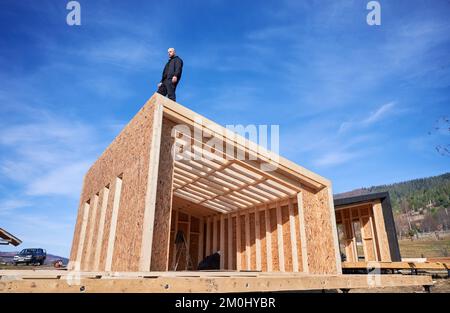 Male builder building wooden frame house. Bald man standing on construction site, inspecting quality of work on sunny day with blue sky on background. Stock Photo