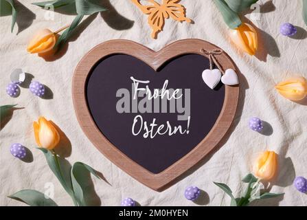 Easter background. Decorated heart blackboard with greeting chalk caption Happy Easter. Orange tulips and quail eggs. Top view, flat lay, Frohe Ostern Stock Photo