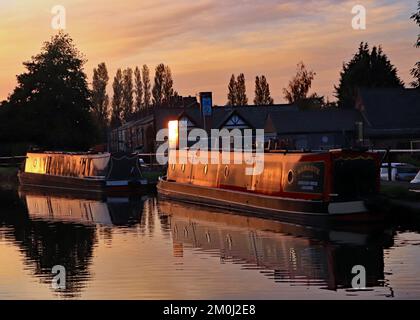 The late afternoon sun is going down as it catches the sides of two Narrowboats moored on the Leeds and Liverpool canal at Burscough in Lancashire. Stock Photo