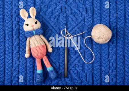 Knitted children's toy hare or rabbit with a ball of thread and a crochet hook on a knitted blue cloth. The symbol of 2023. Stock Photo