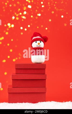 Knitted snowman in a red hat and sweater stands on three red gift boxes on a red background with golden Christmas lights. Merry Christmas and New Year Stock Photo