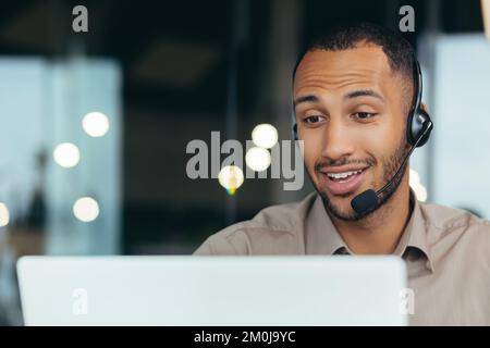 Close-up photo. Learning online. A young handsome African-American student is studying remotely from a laptop, sitting at a desk in a headset, talking on a video call. Stock Photo