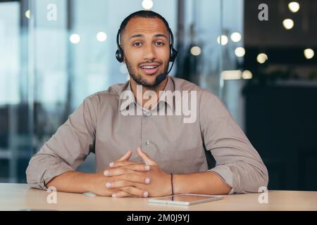 Portrait of a young handsome African American man. He sits in a headset in front of the camera, talks, consults. Business training, webinar, customer support service, online store. Stock Photo