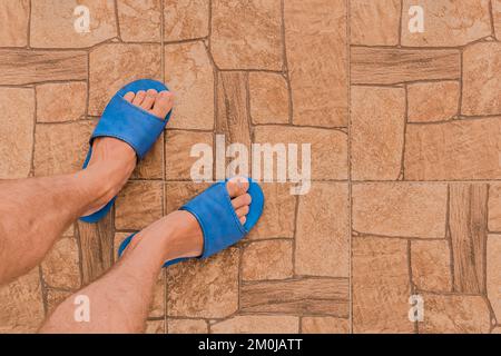 Male feet in blue house slippers stand on brown tile floor with abstract stone pattern texture background, top view.