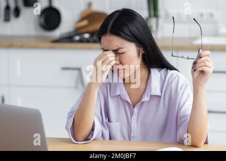 Tired stressed Asian woman freelancer or student having headache rubbing the bridge of her nose Stock Photo