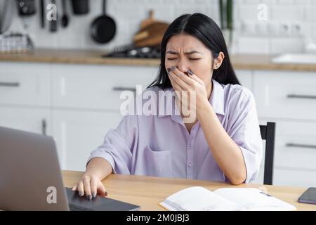 tired Asian woman freelancer or student yawns covers mouth sitting at the workplace at home Stock Photo