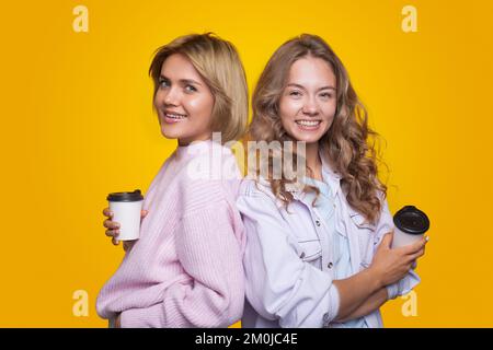 Two blonde girls standing back to back holding a cup of coffee in their hands, looking at the camera. Happy girls friends together. Stock Photo