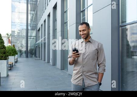Latin american businessman in casual shirt walking near office building from outside, man smiling and using phone, freelancer typing message and browsing internet pages. Stock Photo