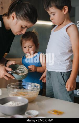 Happy mother and her two little kids making batter for muffins at home kitchen. Sweet food. Happy family concept. Stock Photo