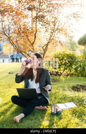 Beautiful woman relaxing drinking a cup of coffee after work hard and using laptop while sitting on green grass in the park. Technology and freedom Stock Photo