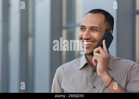 Cheerful and smiling African American office worker talking on the phone, man outside office building in shirt walking in the city, businessman on lunch break. Stock Photo