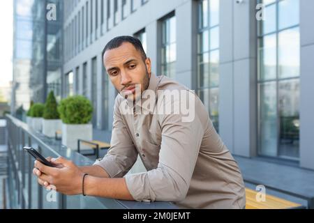 Serious thinking businessman outside office building pondering decision, african american worker sad holding phone in hand. Stock Photo