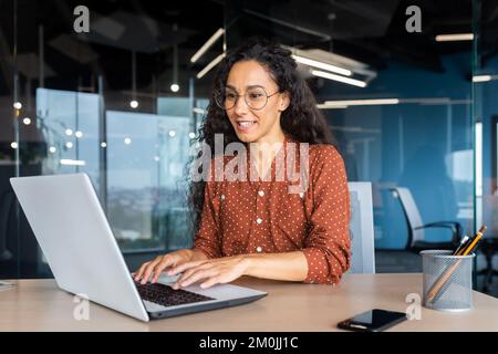 Happy and smiling hispanic businesswoman typing on laptop, office worker with curly hair and glasses happy with achievement results, at work inside office building. Stock Photo