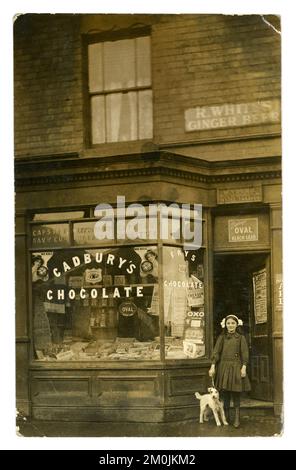 Original pre WW1 postcard of young girl called Gladys, with a pet dog terrier, standing outside her parent's tobacconists /sweet shop front  - W. Young tobacconist. The shop also sells sweets and there's a Cadbury's chocolate sign in the window. The shopkeeper peers through the window. Posters for the silent films in the shop window for 'Out of the Deep' 1912 silent film, 'Buckskin Jack' 1911, Four Singing Miners (named this up to 1912) Photo dated 1912. There's a West Bromwich football fixtures advert which indicates the town was West Bromwich, Staffordshire, West Midlands, England, UK. Stock Photo