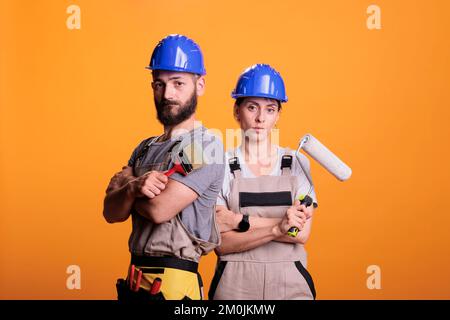 Professional renovators posing with painting tools, standing over yellow background. Team of constructors holding paintbrush and roller brush to work on refurbishment project in studio. Stock Photo