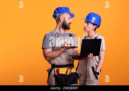 Team of constructors holding laptop and browsing internet online, discussing about renovation project. Construction workers using wireless pc to search building inspiration, studio shot. Stock Photo