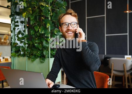 A young red-haired guy, a programmer or entrepreneur in glasses, sits in a stylish cafe at a laptop and speaks on the phone. Freelancer works remotely Stock Photo