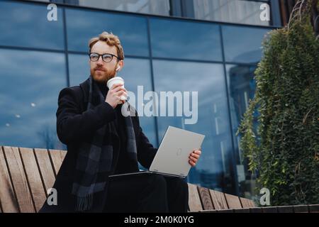 Successful businessman on the background of the business center. Red-haired young guy, in a stylish coat, wireless headphones with a laptop. Stock Photo