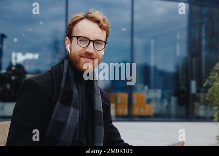 Successful businessman on the background of the business center. Red-haired young guy, in a stylish coat, wireless headphones with a laptop. Stock Photo