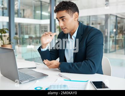 Young serious mixed race businessman talking on a video call using a laptop alone at work. One focused hispanic businessperson talking on a virtual Stock Photo