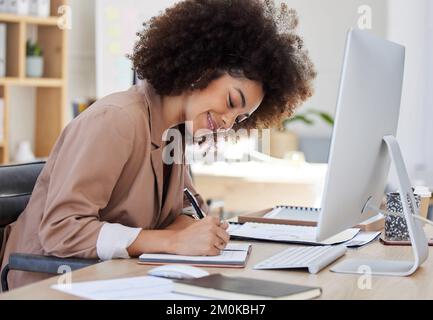 Young smiling mixed race businesswoman with an afro sitting alone in an office and writing notes in a book. Ambitious hispanic creative professional Stock Photo