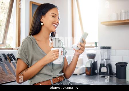 Young mixed race hispanic woman using a phone while drinking coffee in the morning at home. Happy woman using social media on her phone standing in Stock Photo