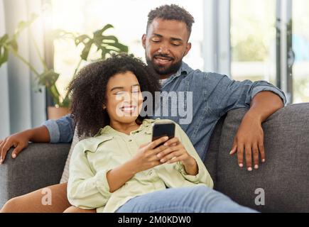 Happy mixed race couple smiling while using a phone together at home. Carefree loving hispanic boyfriend and girlfriend bonding and using social media Stock Photo