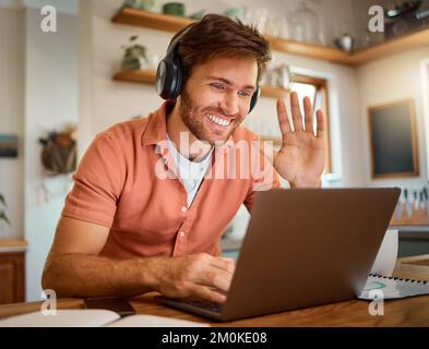 Young happy caucasian businessman wearing headphones and waving his hand on a virtual meeting on a laptop at home alone. One joyful male Stock Photo