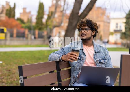 Man looking at the camera in the park with a laptop, drinking coffee on a background of green trees, a hot sunny summer day. Stock Photo
