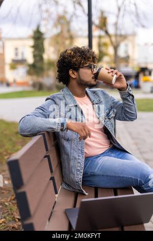 Indian man sitting on a park bench and drinking coffee while working on laptop in outdoor fresh air at day time. Stock Photo