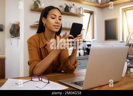 Young mixed race woman drinking a cup of coffee while using her phone and working on a laptop alone in the morning in the kitchen. Content hispanic Stock Photo