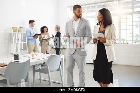 Mixed race businessman and african american businesswoman talking while using a digital tablet in an office with coworkers. Businesspeople working Stock Photo