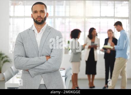 Young mixed race businessman standing with arms crossed while in an office with colleagues. Serious hispanic male boss In a meeting with coworkers Stock Photo