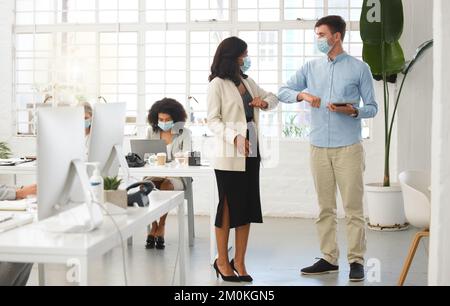 Diverse colleagues talking and greeting each other with their elbows while wearing masks to protect from infection of a virus. Young caucasian Stock Photo